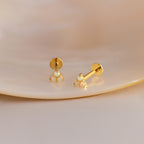 A close-up photograph displays a pair of dainty gold stud earrings resting on a smooth, neutral-toned ceramic surface. Each earring features a triangular cluster of three tiny round opals, each held in place with polished gold prongs. The iridescent opals reflect hues of green, blue, and pink, subtly catching the light.