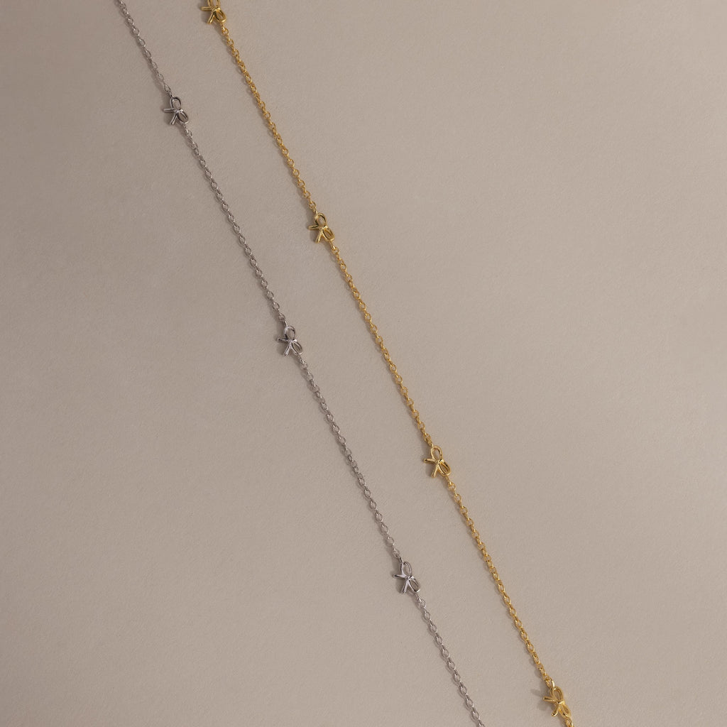 Side-by-side flat-lay of the Dainty Bow Necklace in both gold and silver finishes on a soft beige background.