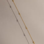 Side-by-side flat-lay of the Dainty Bow Necklace in both gold and silver finishes on a soft beige background.