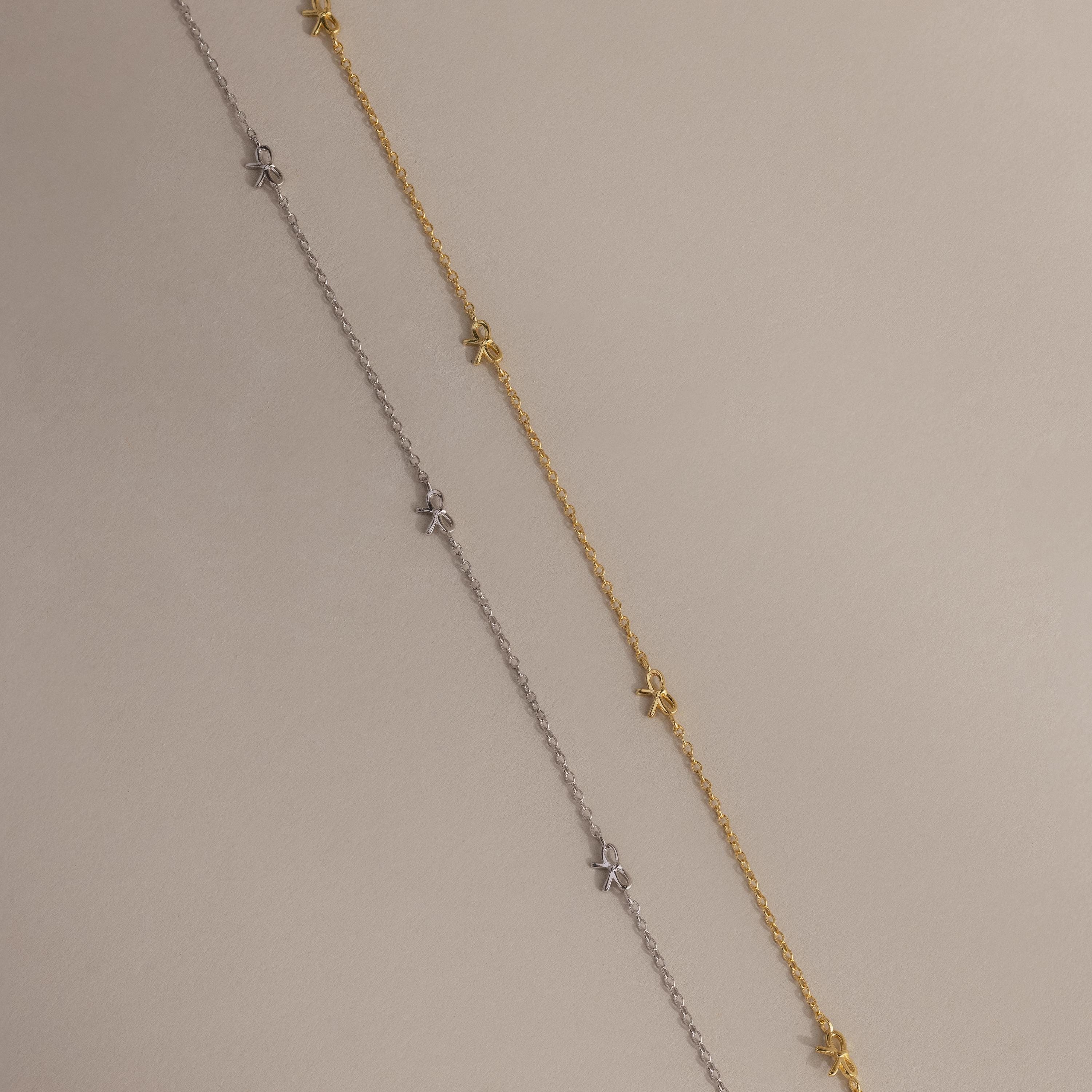 Side-by-side flat-lay of the Dainty Bow Necklace in both gold and silver finishes on a soft beige background.
