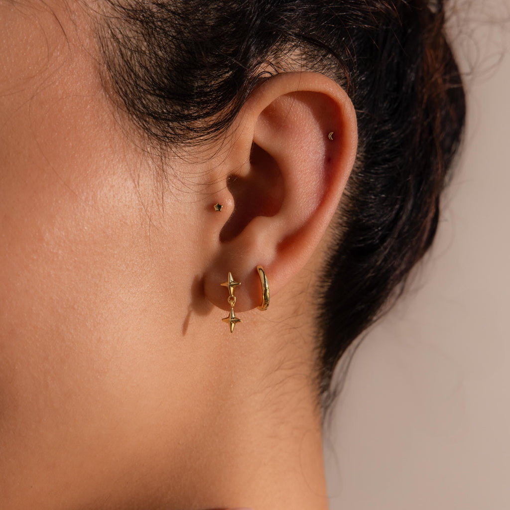 A close-up of a woman's ear adorned with multiple gold earrings, featuring the double star drop flat back earring in the lower lobe and a small hoop above.