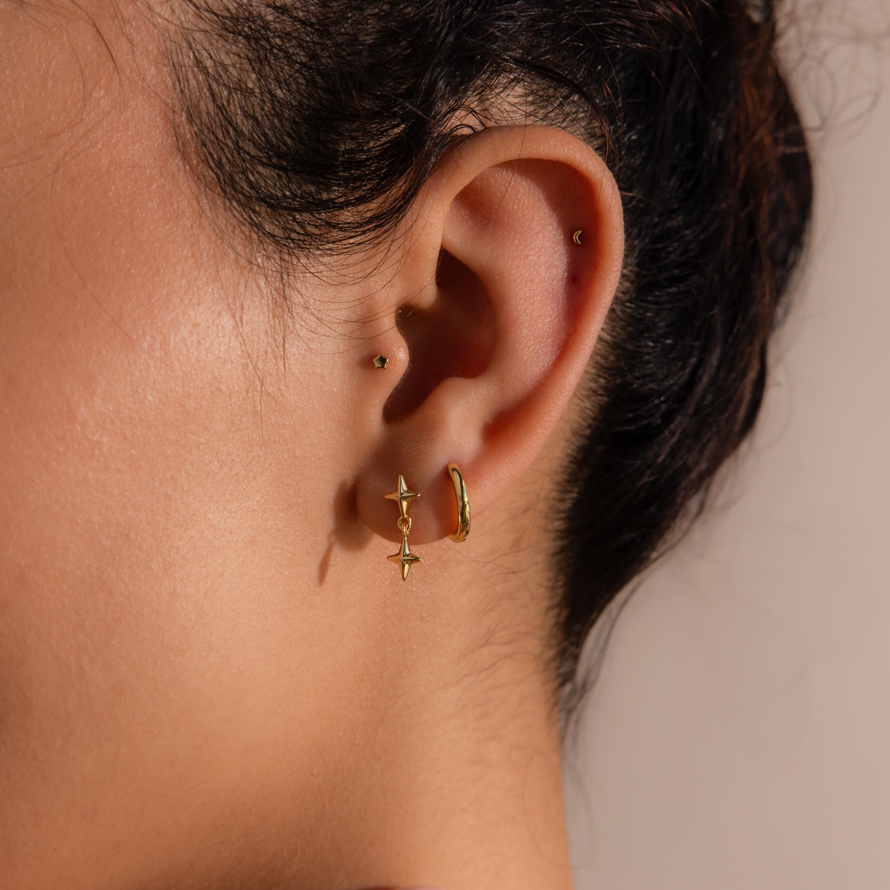 A close-up of a woman's ear adorned with multiple gold earrings, featuring the double star drop flat back earring in the lower lobe and a small hoop above.