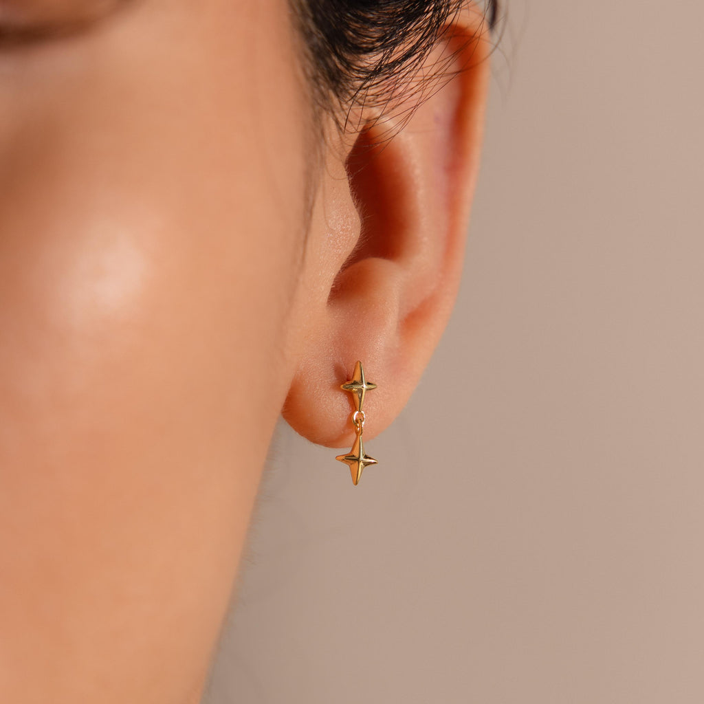 A close-up view of a woman's ear adorned with a gold flat back earring featuring two vertically aligned, four-pointed star charms. The top star sits snugly against the earlobe, while the second star dangles just below, catching the light with a polished gold finish.