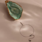 Flat lay of a dainty silver hand chain bracelet displayed on a soft beige surface, with the chain draped across a green and gold oyster shell. The bracelet features three bezel-set diamonds and a heart charm at the lobster clasp.