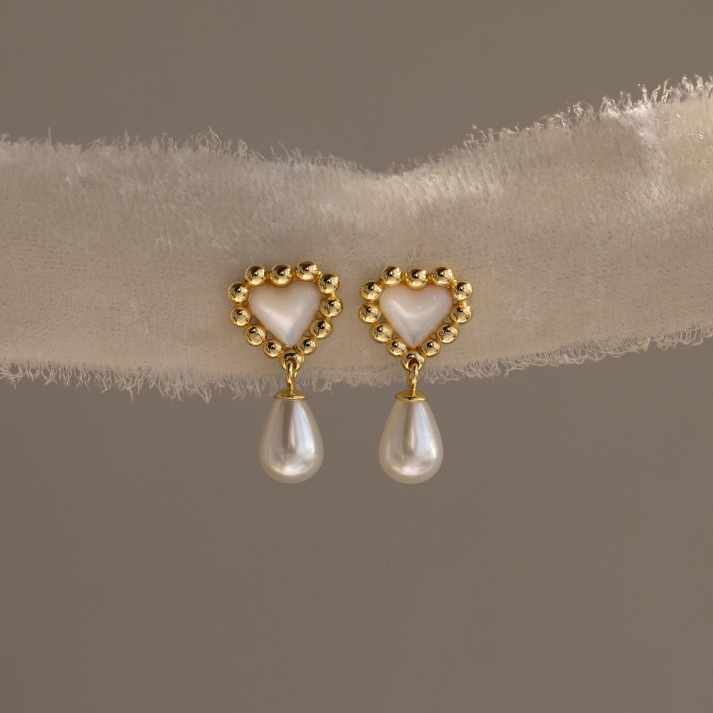 Pair of gold beaded heart earrings with mother-of-pearl centers and dangling pearls displayed on a soft textured fabric.