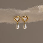 Pair of gold beaded heart earrings with mother-of-pearl centers and dangling pearls displayed on a soft textured fabric.