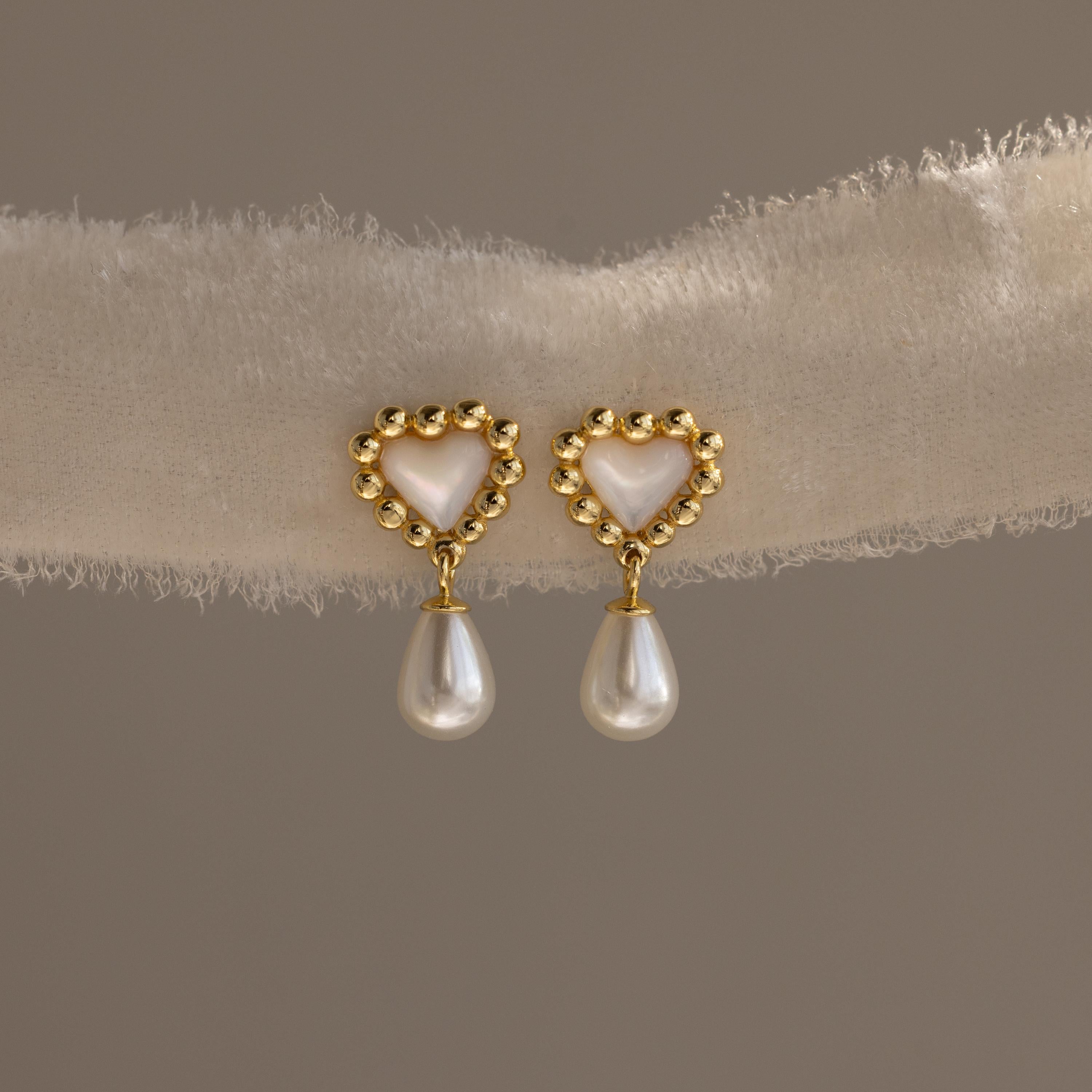 Pair of gold beaded heart earrings with mother-of-pearl centers and dangling pearls displayed on a soft textured fabric.