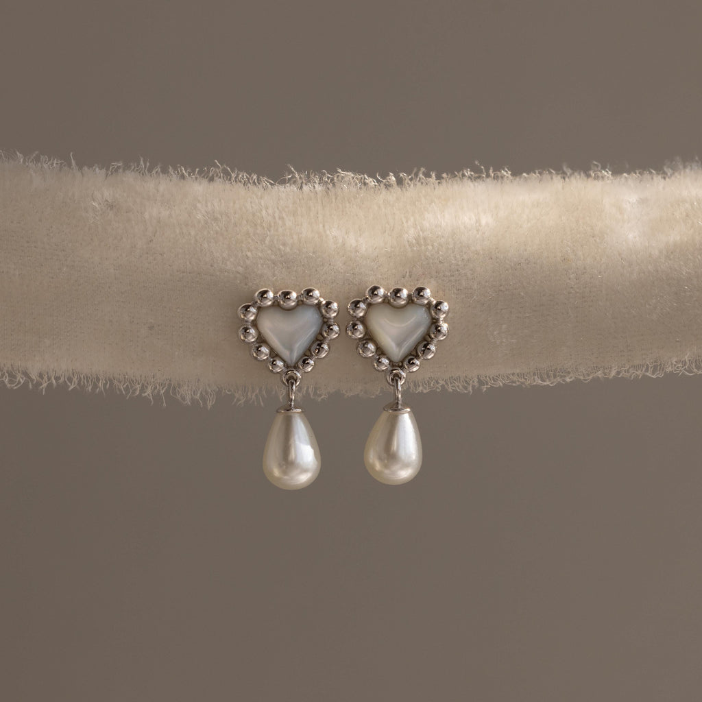 Silver heart earrings with beaded edges and pearl drops displayed on a soft neutral-toned fabric roll.
