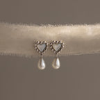 Silver heart earrings with beaded edges and pearl drops displayed on a soft neutral-toned fabric roll.