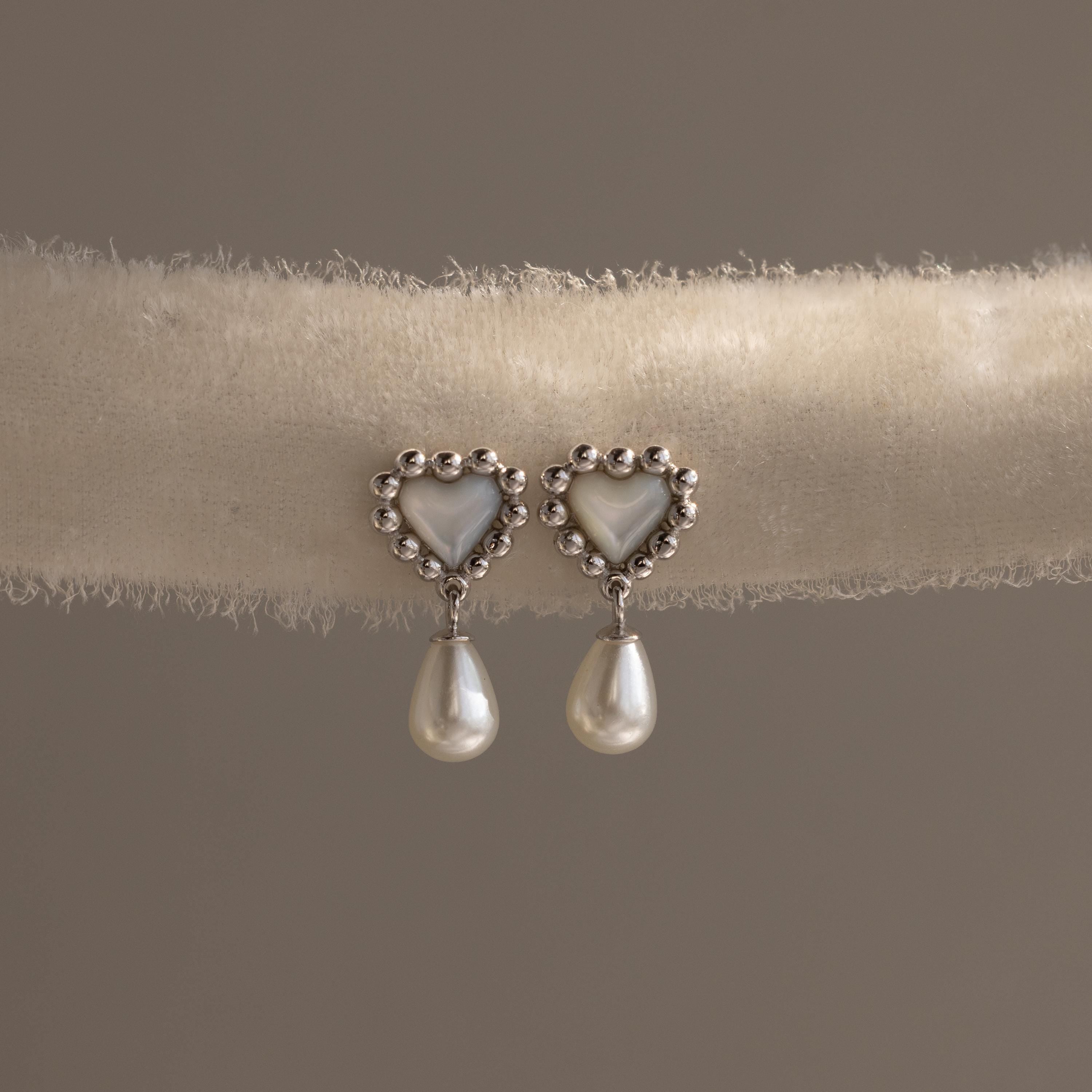 Silver heart earrings with beaded edges and pearl drops displayed on a soft neutral-toned fabric roll.