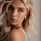 Model with long blonde hair wearing gold beaded pearl heart earrings, looking over shoulder in soft lighting.