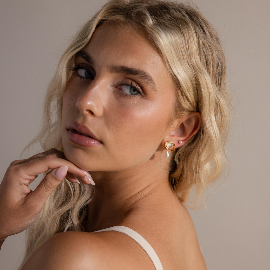 Model wears gold beaded pearl heart drop earrings, stacked with a pearl stud and simple gold huggie hoop