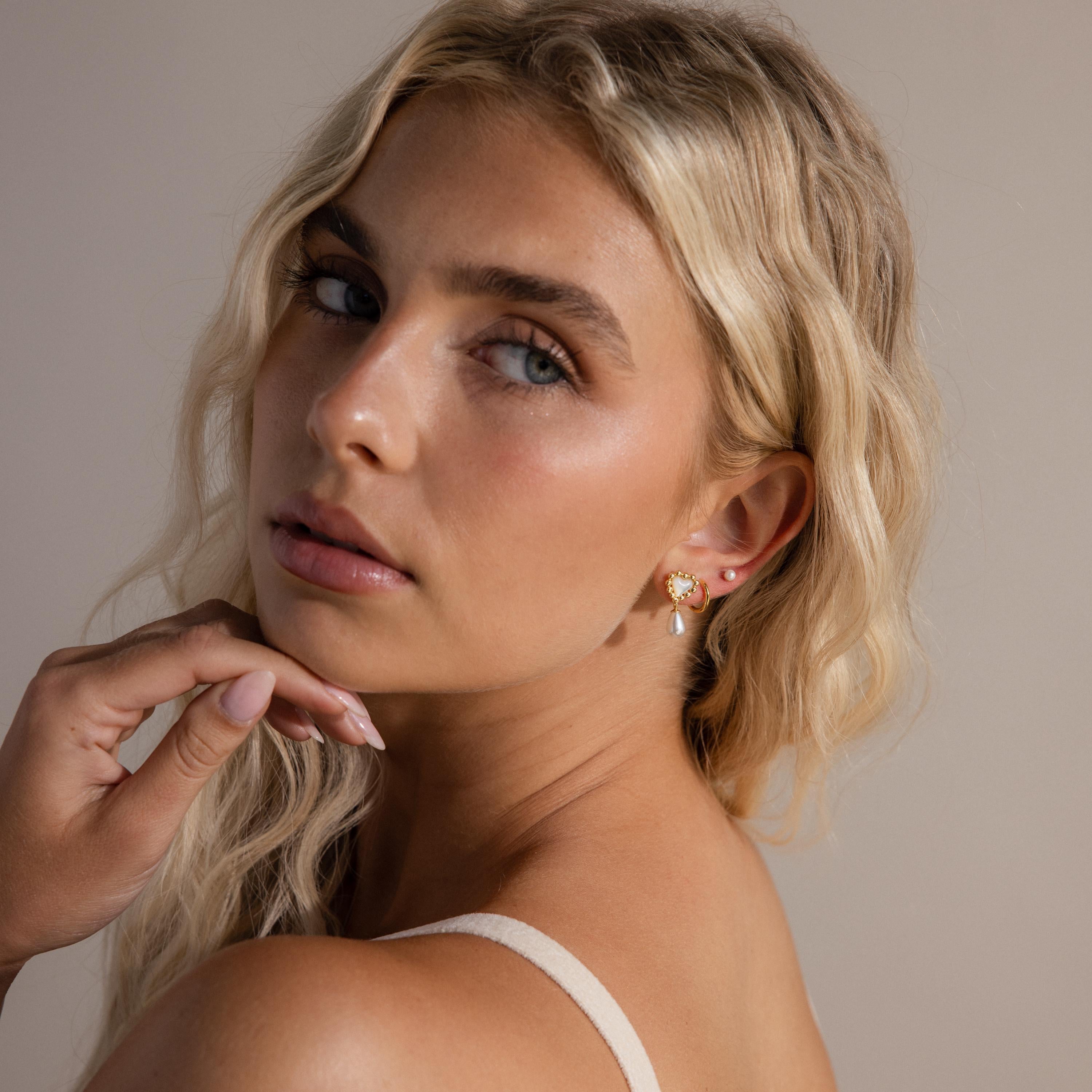 Model wears gold beaded pearl heart drop earrings, stacked with a pearl stud and simple gold huggie hoop