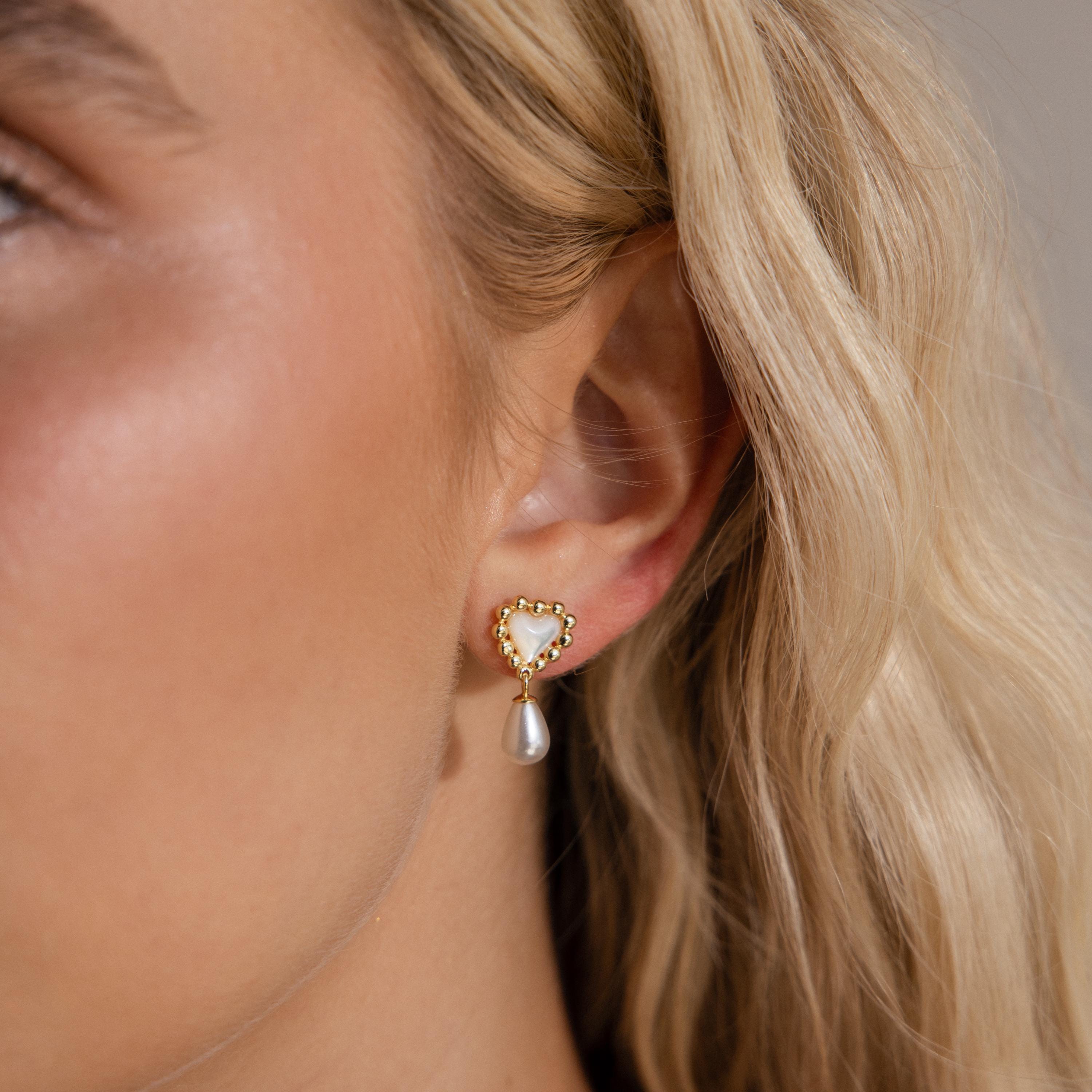 Model wearing gold beaded heart-shaped pearl drop earrings; the earrings feature a mother-of-pearl heart bordered by small gold beads and a dangling teardrop pearl.
