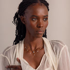 Model with long braided hair wears a sheer cream blouse and two layered gold necklaces. The top necklace features a single marquise-shaped gemstone pendant, while the lower necklace displays a row of small teardrop diamond charms evenly spaced along the chain. She gazes slightly to the side with a serene, composed expression.