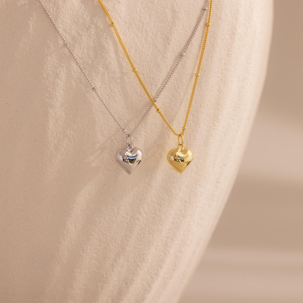 Both the 18k gold and white gold heart necklaces are displayed hanging next to each other against a pale, curved backdrop