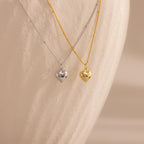 Both the 18k gold and white gold heart necklaces are displayed hanging next to each other against a pale, curved backdrop