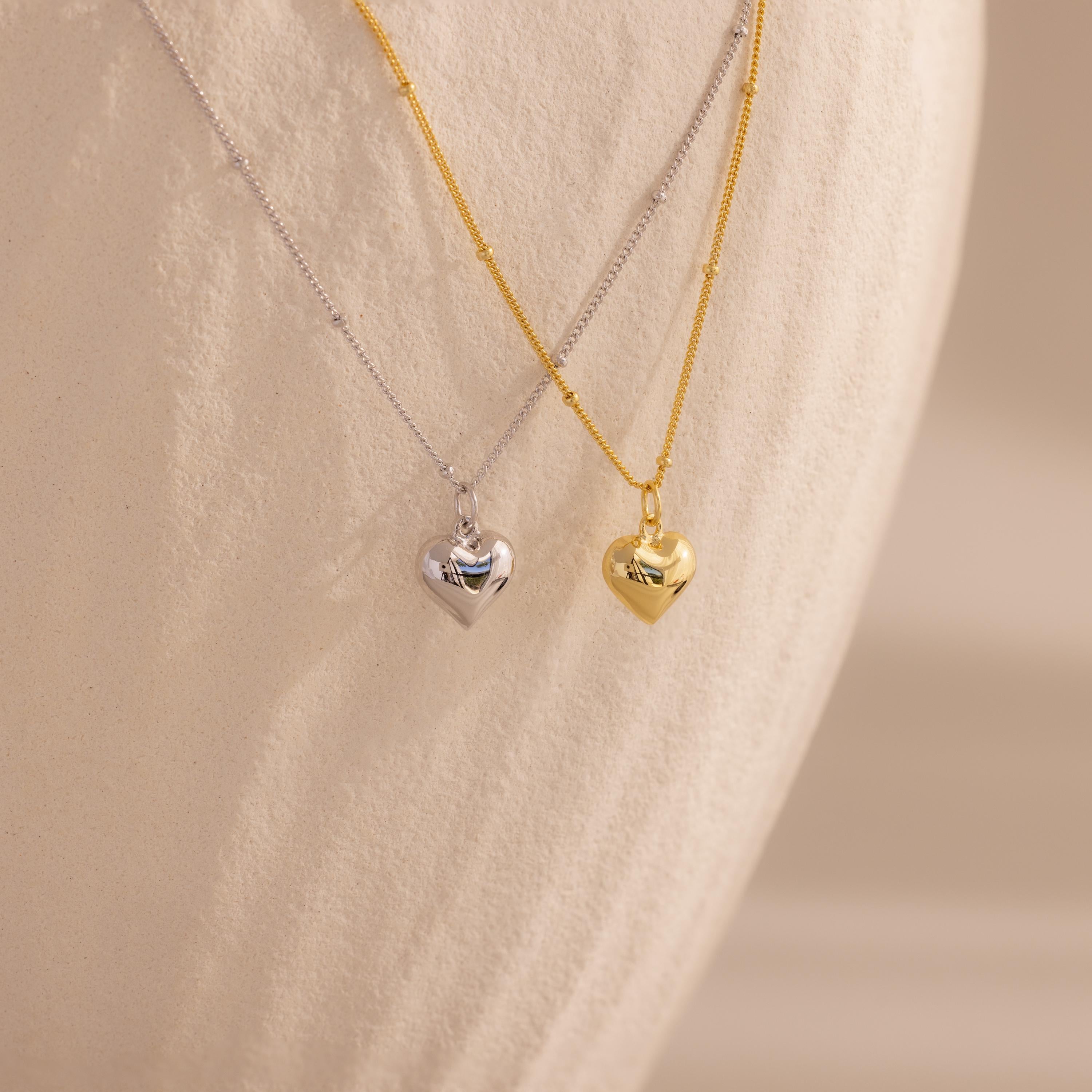 Both the 18k gold and white gold heart necklaces are displayed hanging next to each other against a pale, curved backdrop