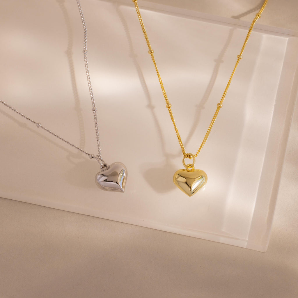Side-by-side product shot of both colorways: the 18k gold and white gold Puff Heart Pendants rest on a translucent display surface