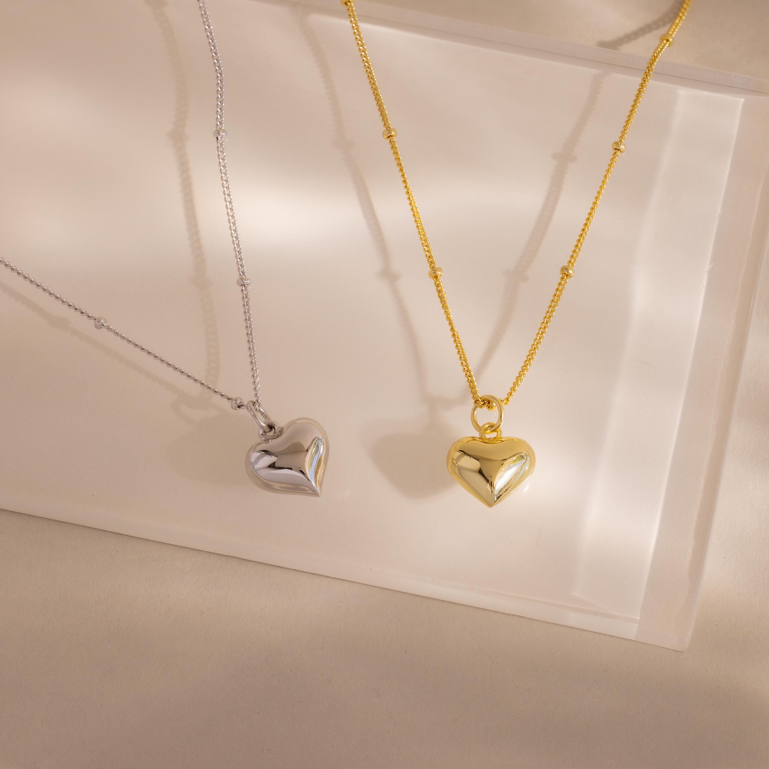 Side-by-side product shot of both colorways: the 18k gold and white gold Puff Heart Pendants rest on a translucent display surface