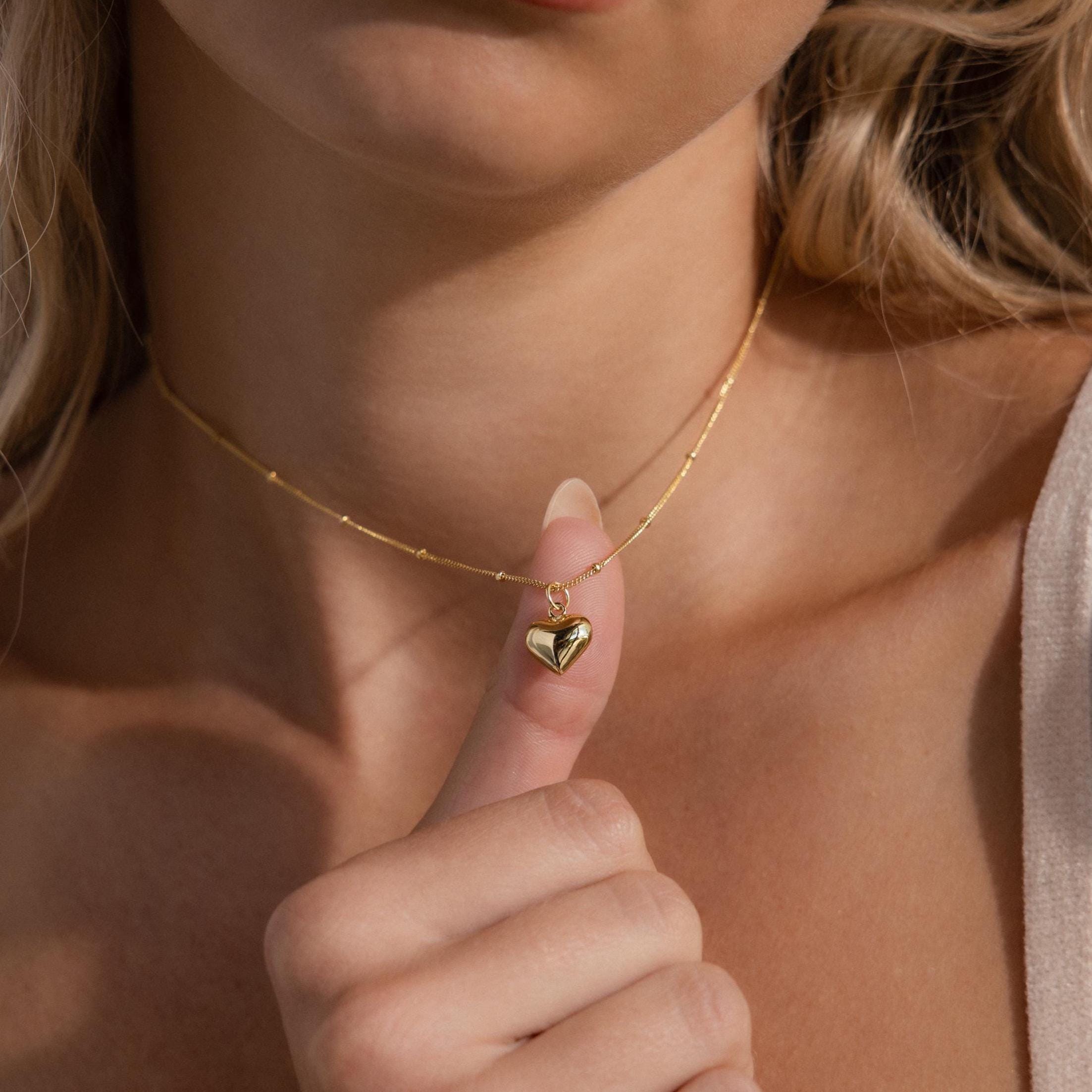 A close-up shot of a woman’s neck and chest as she gently holds a dainty gold heart pendant between her thumb and index finger, drawing attention to its glossy, puffed silhouette. The pendant is suspended on a satellite 18k gold chain that drapes delicately around her collarbone,