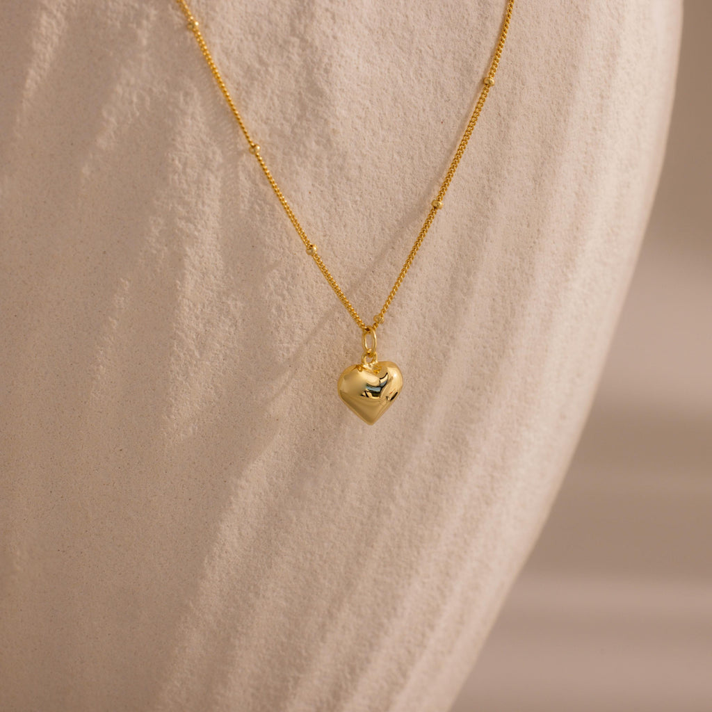 Gold Puff Heart Pendant Necklace displayed against a textured, ivory-toned background.
