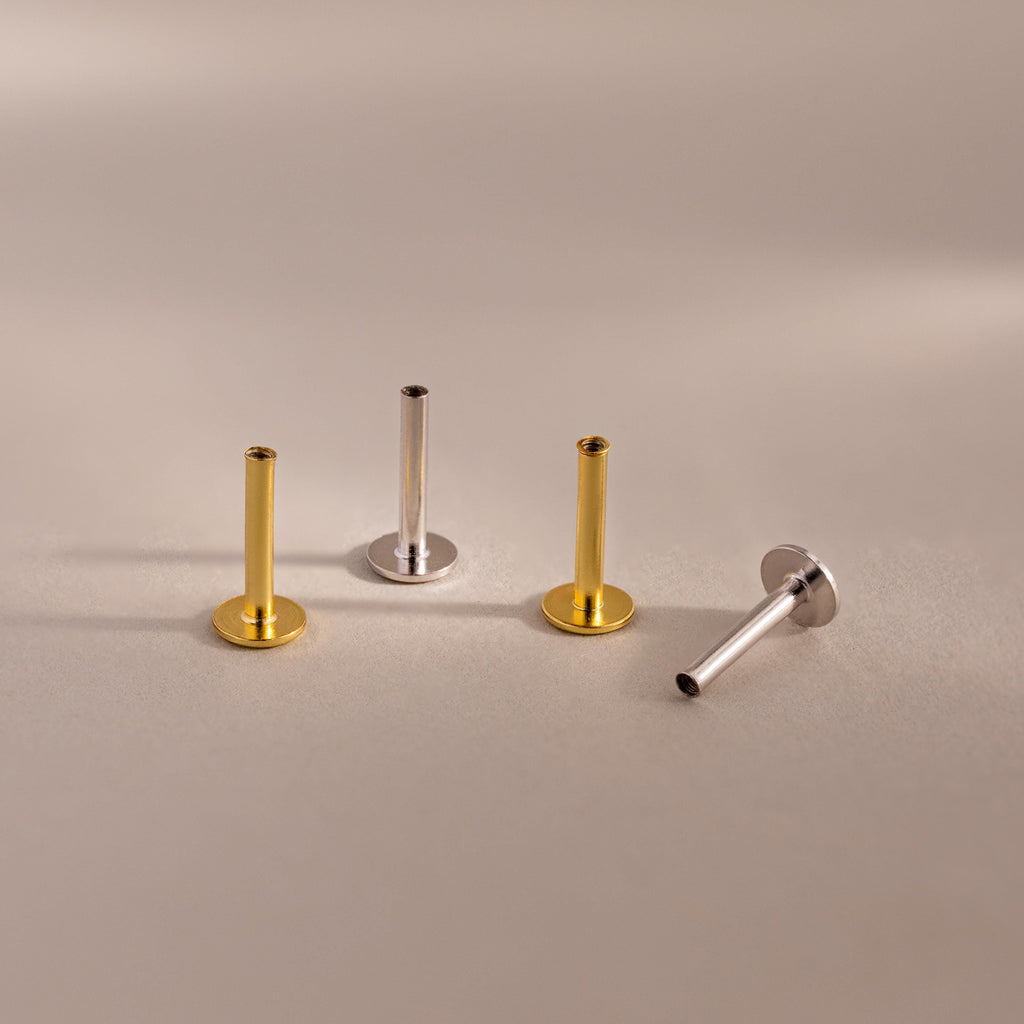 A mixed set of four Flat Back Posts — two 18k gold and two sterling silver — arranged upright and diagonally on a beige background.