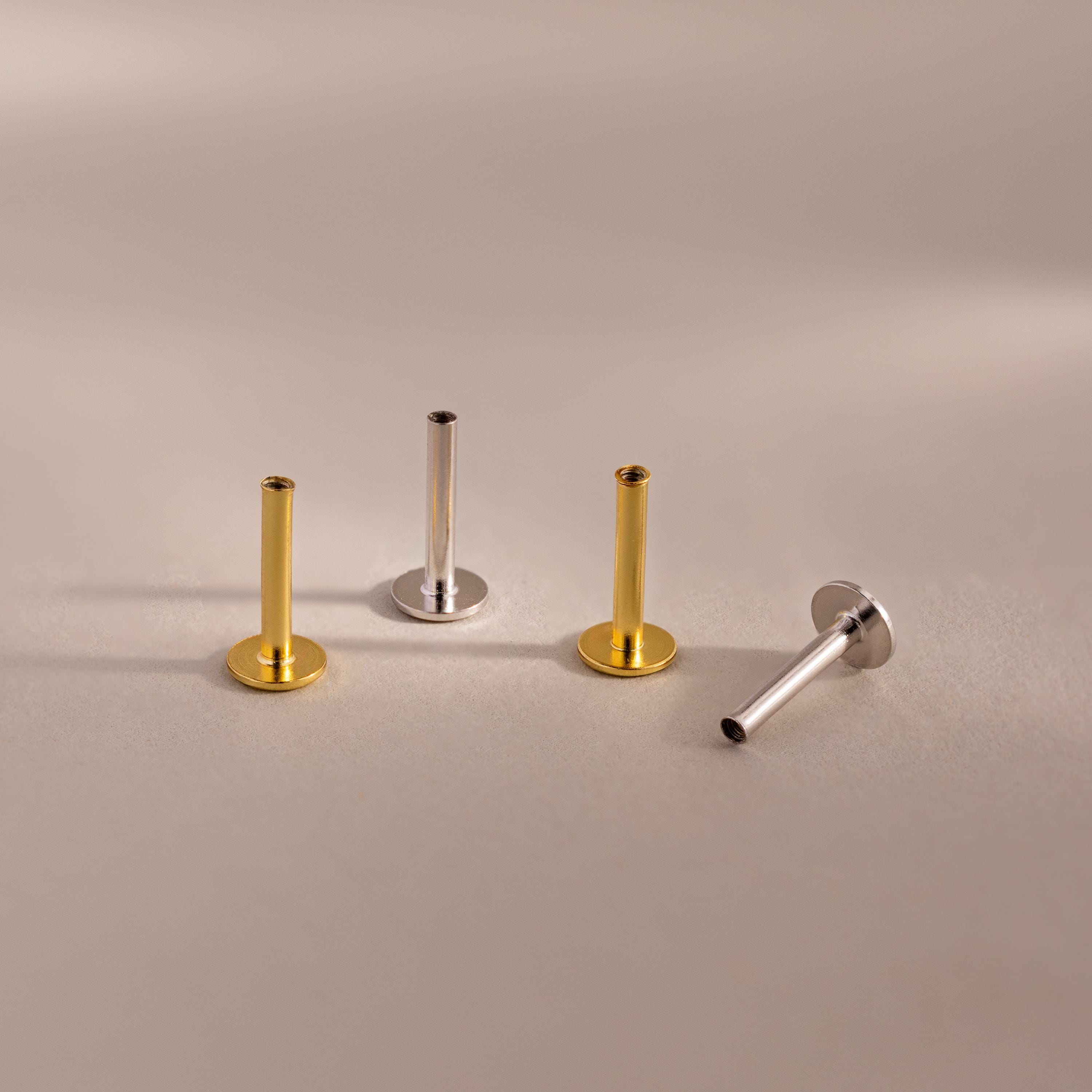 A mixed set of four Flat Back Posts — two 18k gold and two sterling silver — arranged upright and diagonally on a beige background.