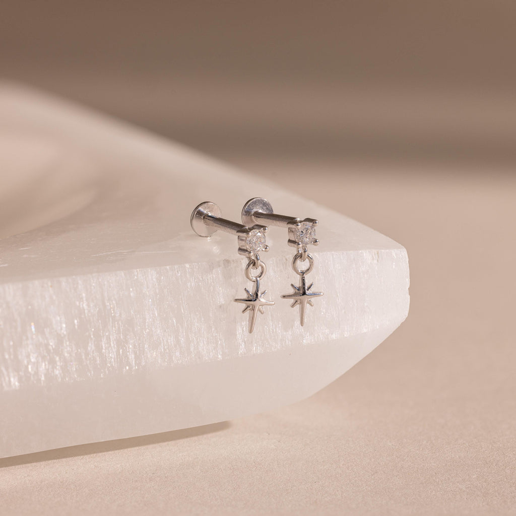 Pair of silver flatback earrings featuring a small round diamond stud and dangling star charms, displayed on a textured white crystal surface.