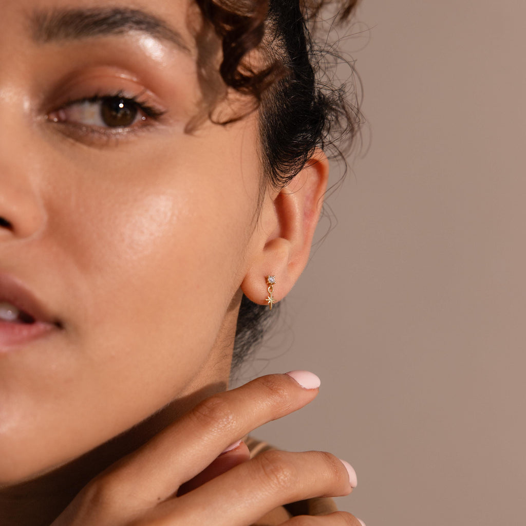 Close-up of a woman wearing a gold flatback earring with a small diamond stud and dangling star charm. Her skin glows, and she gently touches her jawline with soft pink-manicured fingers.