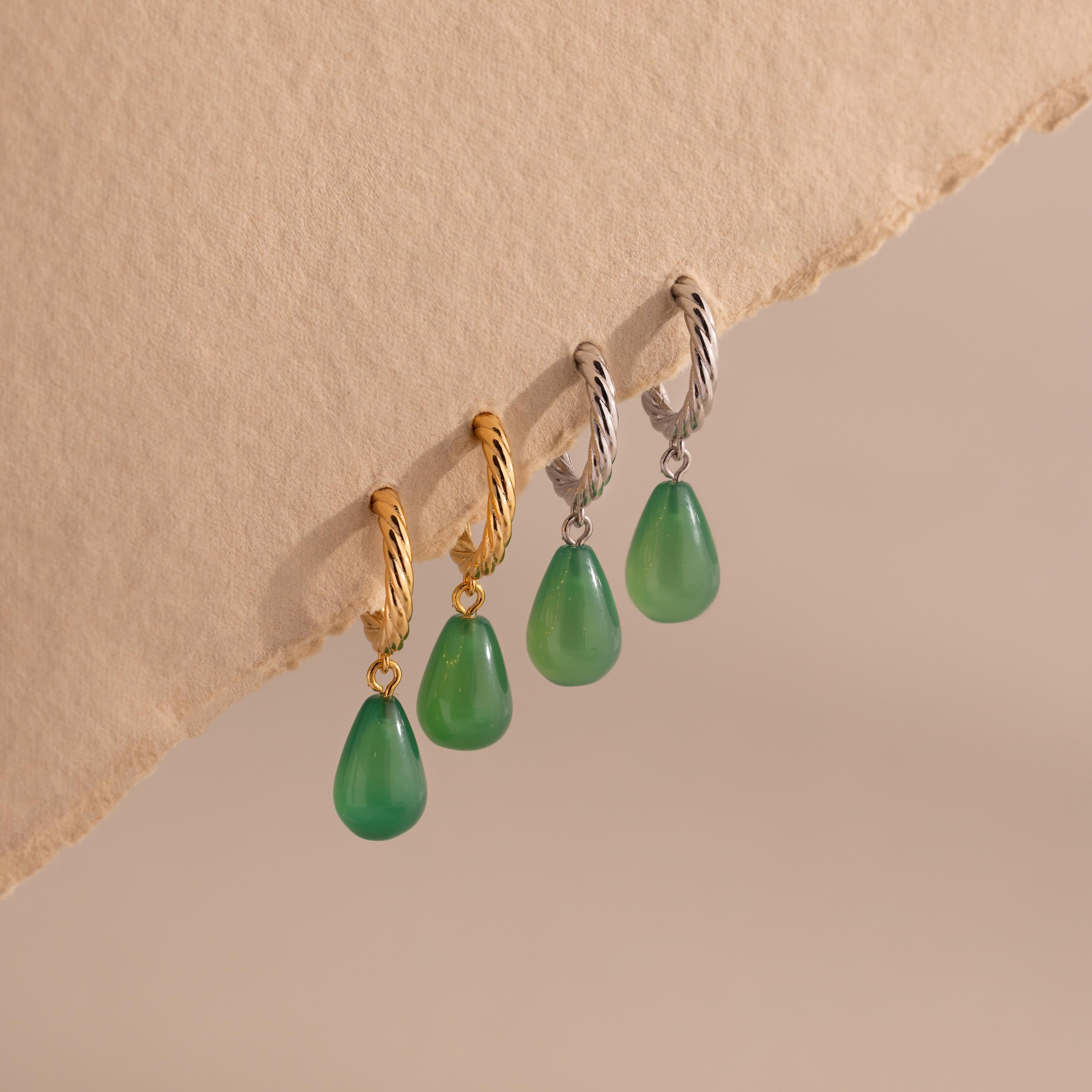 Four twisted hoop earrings with teardrop-shaped green jade charms, displayed in gold and silver finishes on the edge of a textured beige surface.
