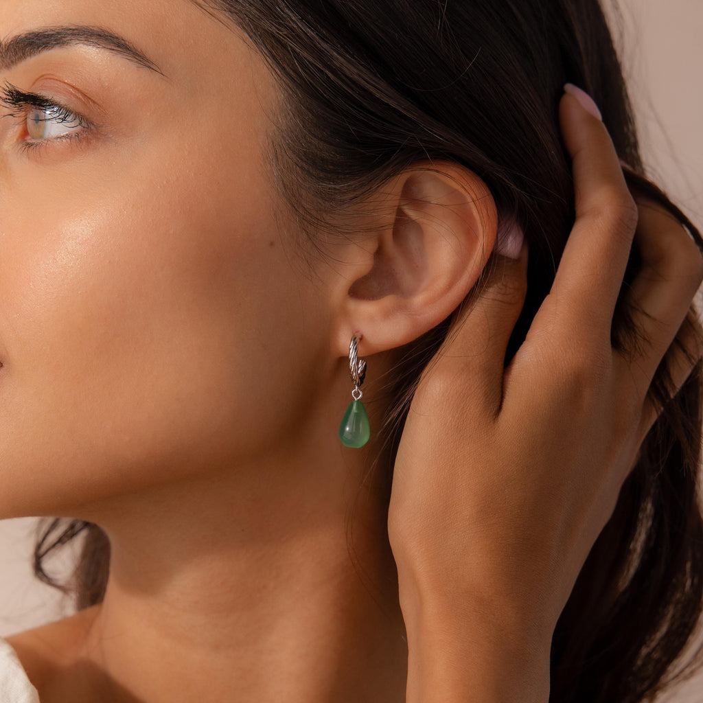 Close-up of a woman wearing a silver twisted hoop earring with a green jade teardrop charm, gently tucking her hair behind her ear.