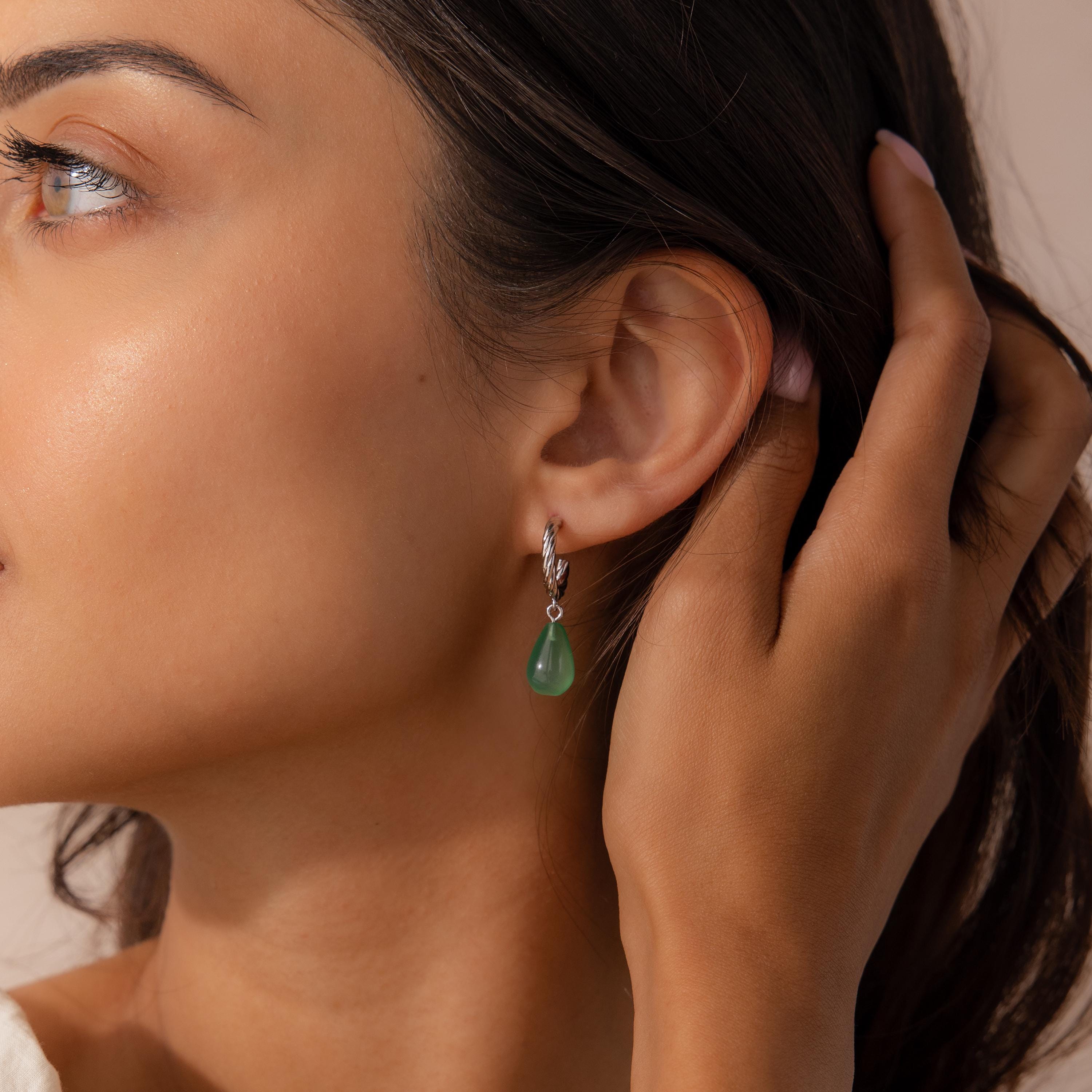 Close-up of a woman wearing a silver twisted hoop earring with a green jade teardrop charm, gently tucking her hair behind her ear.
