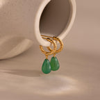Pair of twisted gold hoop earrings with green jade teardrop charms displayed on a smooth beige background.