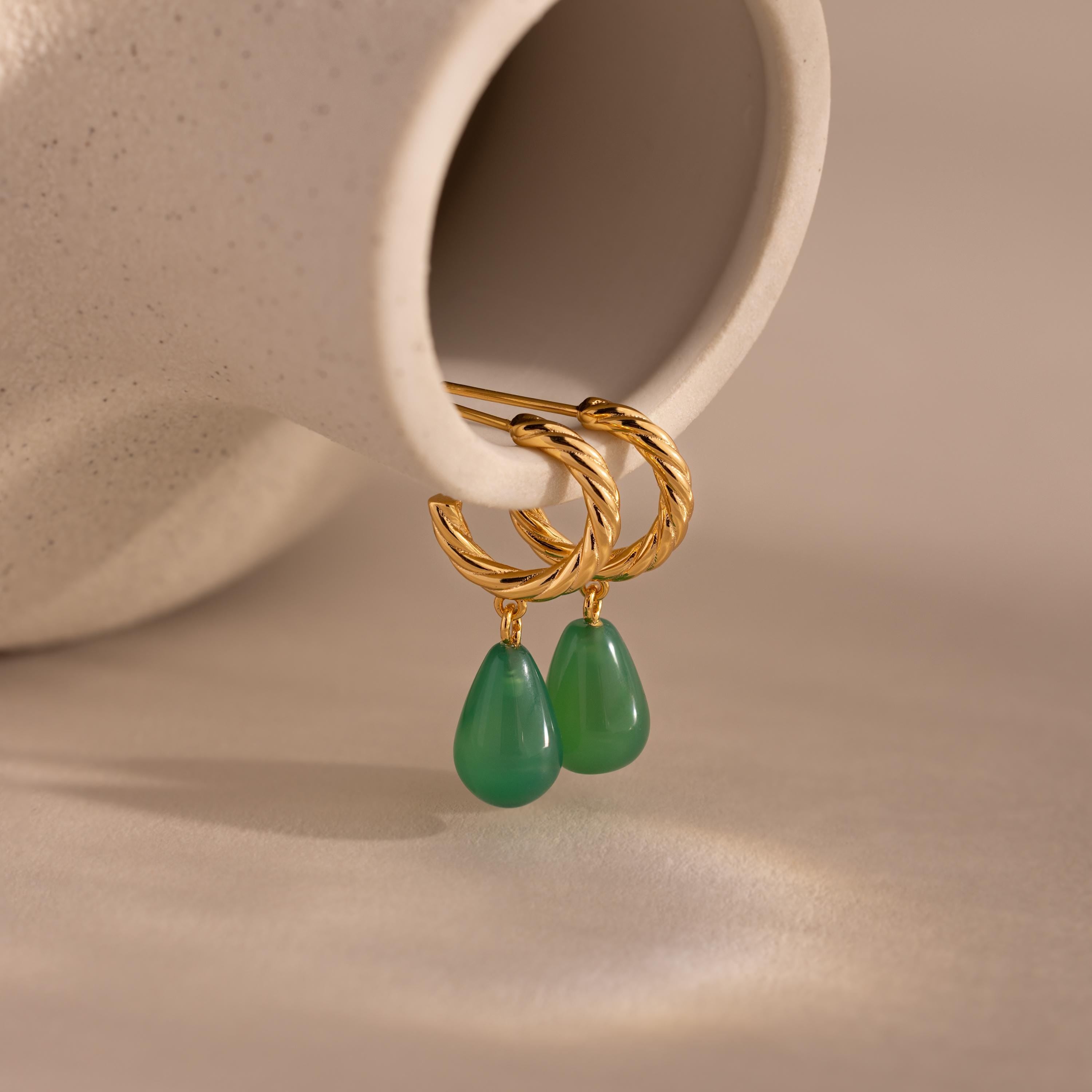 Pair of twisted gold hoop earrings with green jade teardrop charms displayed on a smooth beige background.