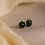 Green jade rose stud earrings with center stones placed on a soft beige surface, partially framed by draped fabric.