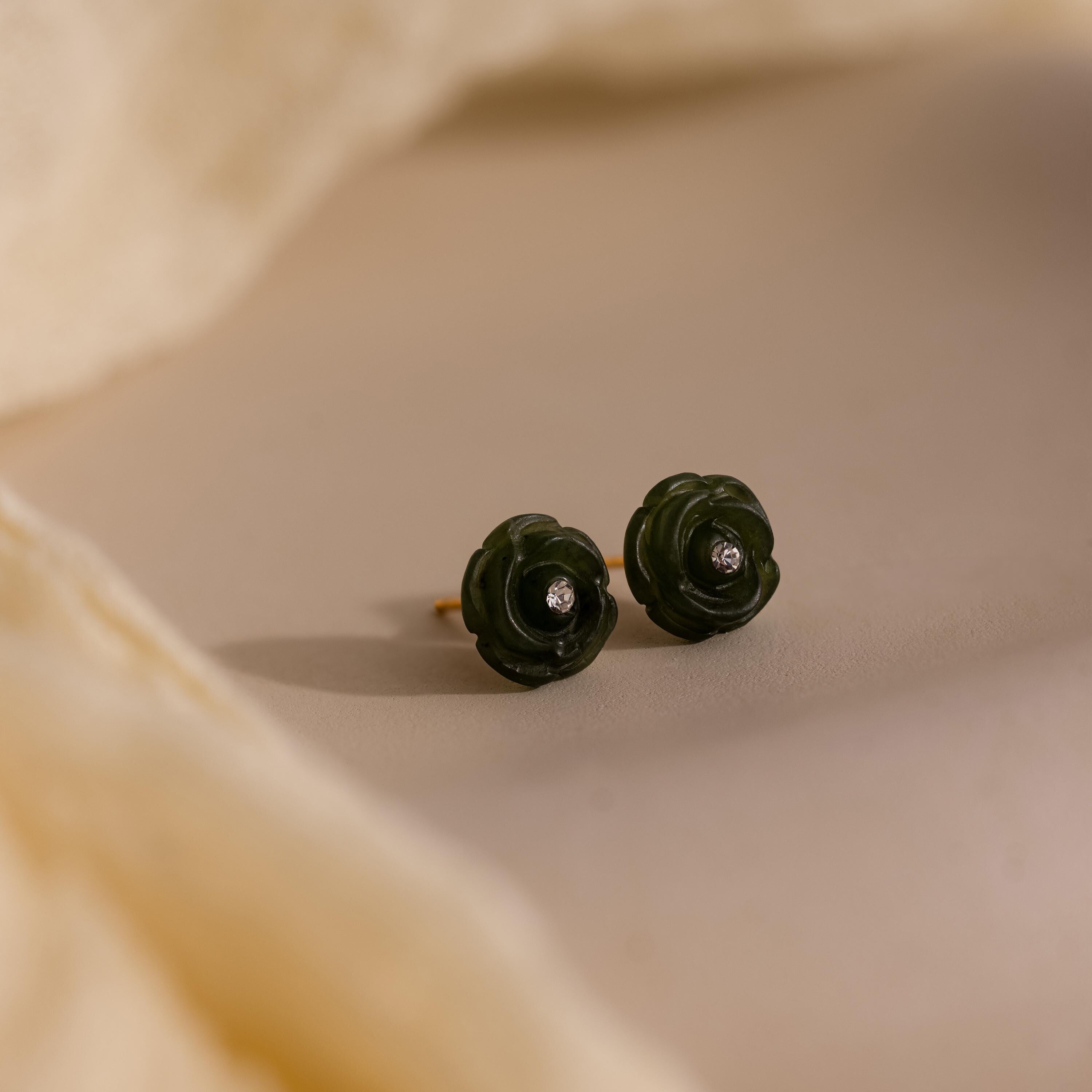 Green jade rose stud earrings with center stones placed on a soft beige surface, partially framed by draped fabric.