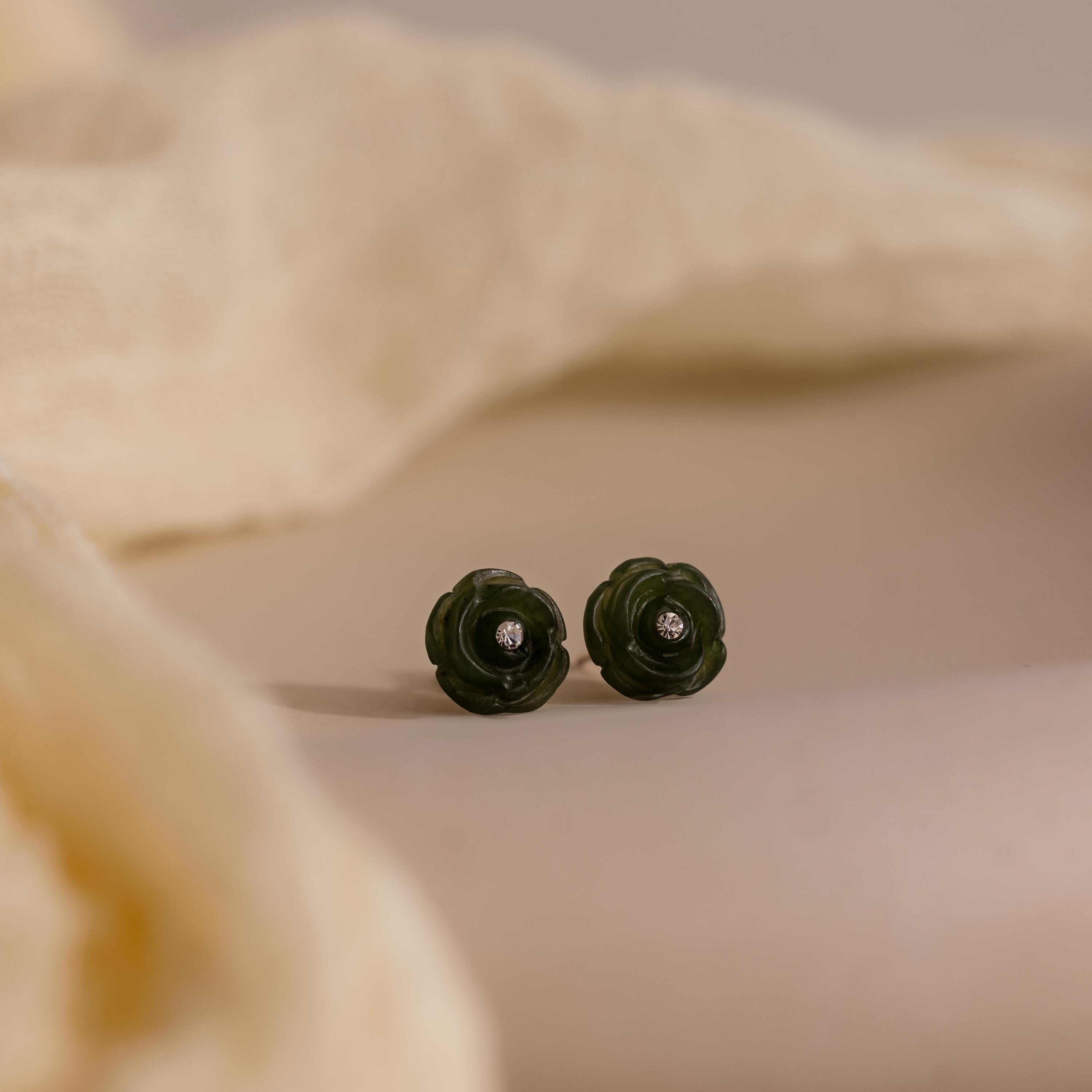 Green jade rose stud earrings with center stones placed on a soft beige surface, partially framed by draped fabric.
