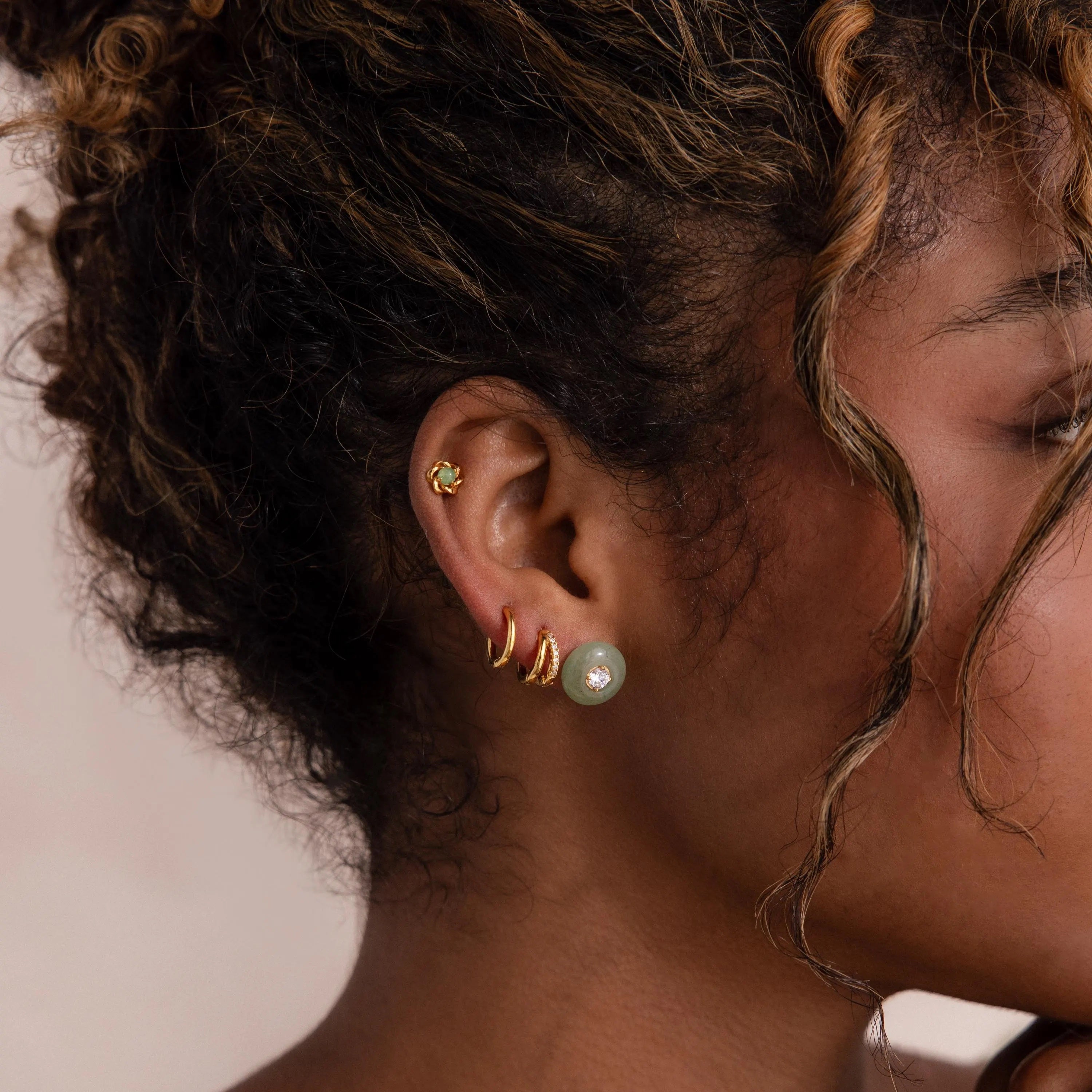 Model wearing a gold flower-shaped jade stud earring in her cartilage, stacked with a round jade earring, and multiple gold huggie hoops.
