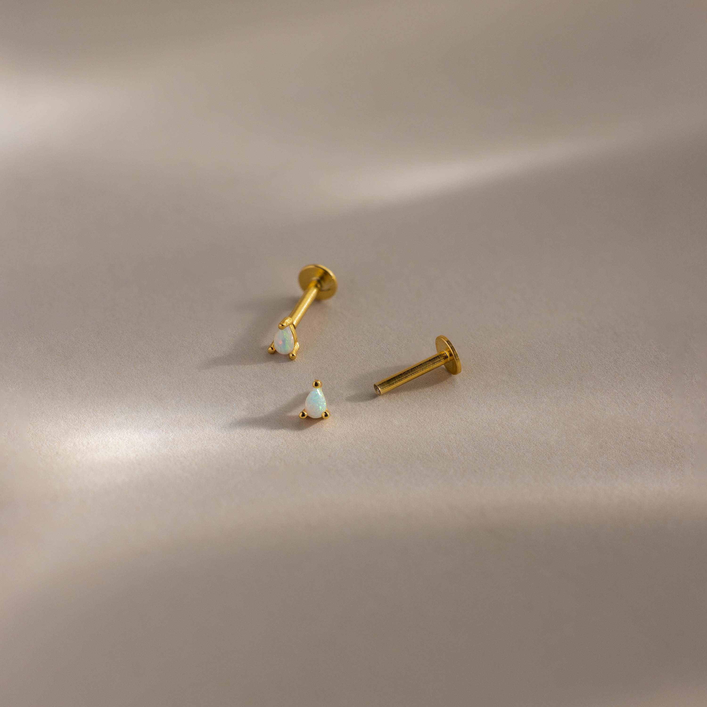 Two gold pear opal studs shown disassembled to reveal the flatback threading design on a minimalist beige surface.