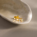 A close-up photograph of a pair of gold pear opal flatback studs resting elegantly inside a glossy, off-white seashell dish. Each stud features a luminous teardrop-shaped opal gemstone set  with three tiny prongs,  capturing hints of pink, green, and blue in the light