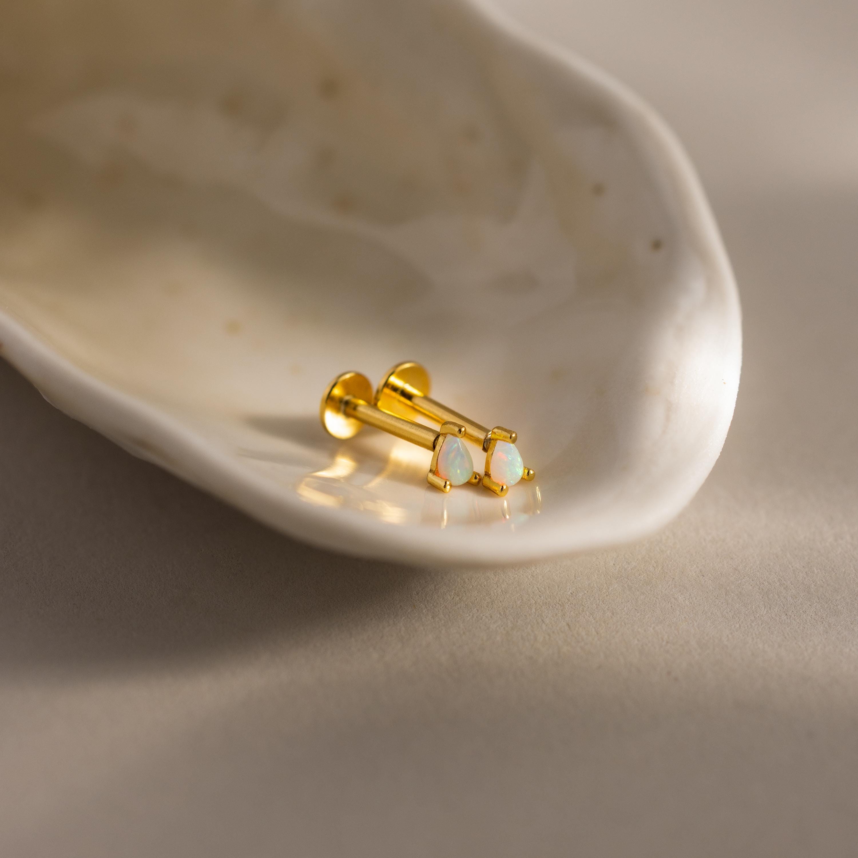 A close-up photograph of a pair of gold pear opal flatback studs resting elegantly inside a glossy, off-white seashell dish. Each stud features a luminous teardrop-shaped opal gemstone set  with three tiny prongs,  capturing hints of pink, green, and blue in the light