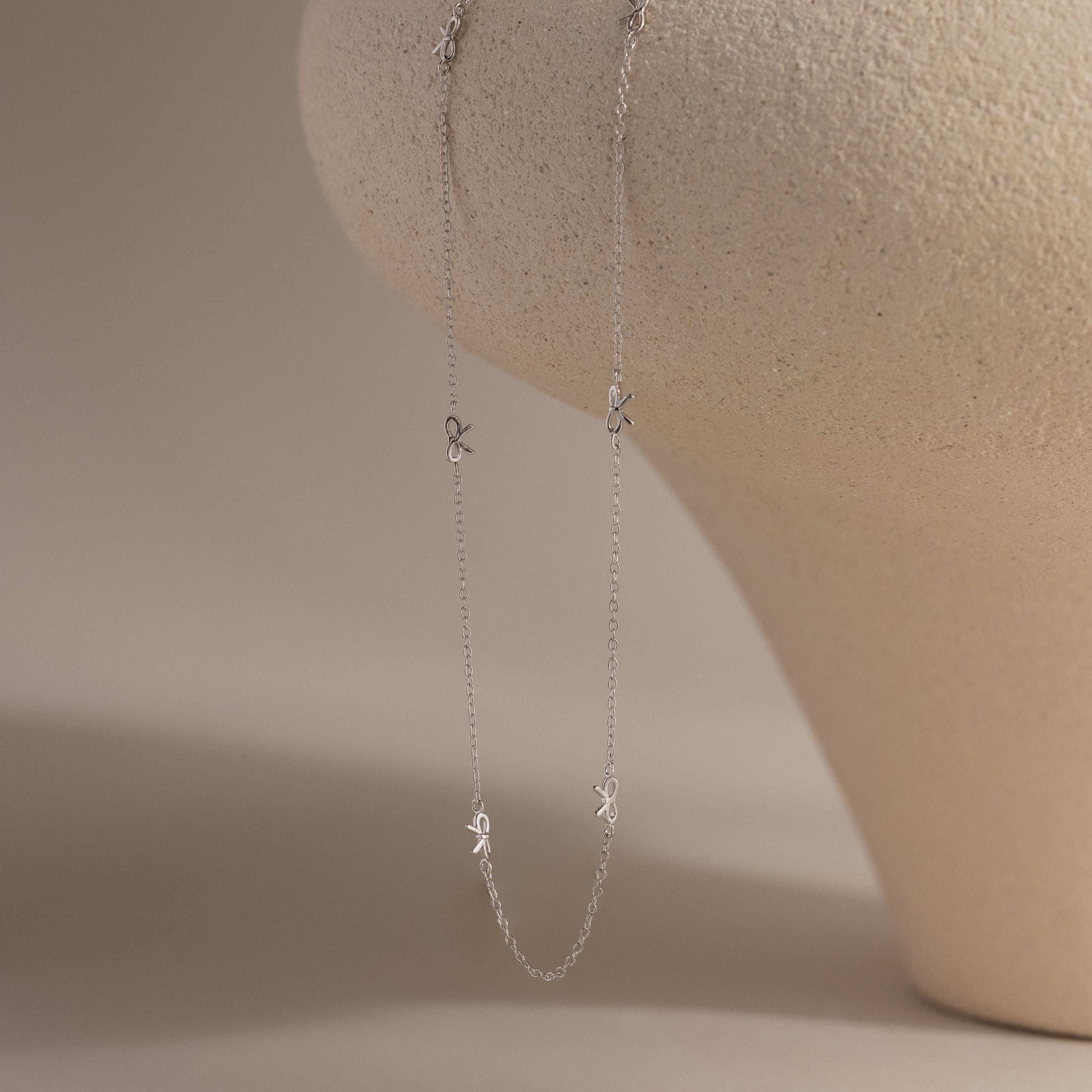 Silver version of the Dainty Bow Necklace displayed hanging on a sculpted neutral-toned jewelry form.