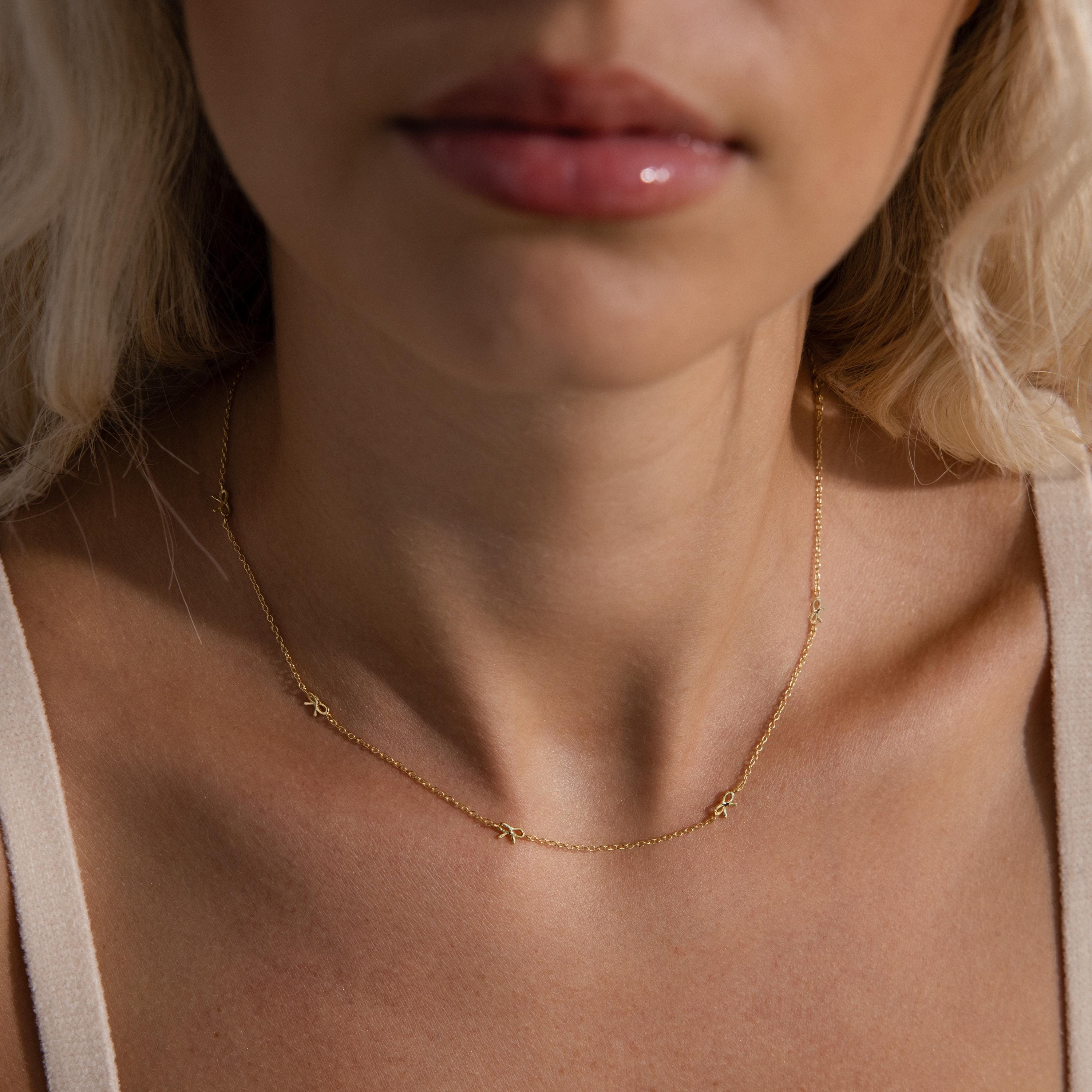 A close-up view of a woman wearing the Dainty Bow Necklace, featuring a fine gold chain adorned with tiny, delicately crafted bow-shaped charms spaced evenly along the collarbone. The necklace’s minimalist yet elegant design catches the light subtly, complementing her natural skin tone and soft styling.