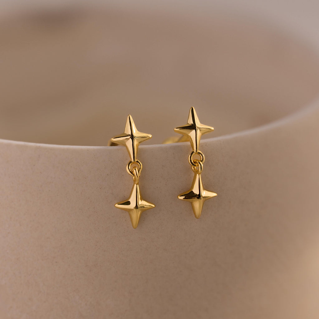 A pair of gold flat back earrings with double star-shaped drops displayed on a curved beige ceramic surface