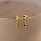 A pair of gold flat back earrings with double star-shaped drops displayed on a curved beige ceramic surface