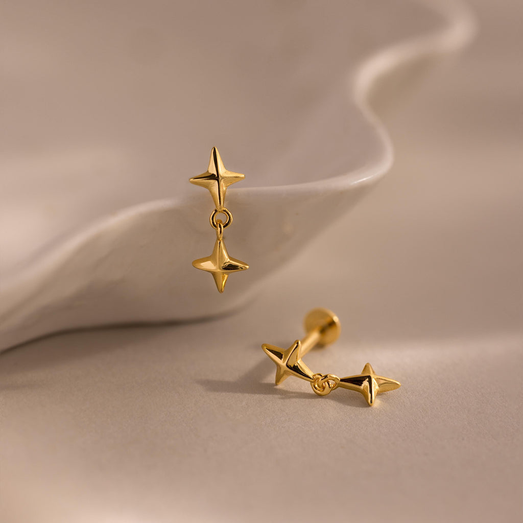 A pair of gold star drop flat back earrings resting against a white ceramic shell dish, with one earring standing upright and the other lying flat, illuminated by soft ambient light.