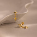 A pair of gold star drop flat back earrings resting against a white ceramic shell dish, with one earring standing upright and the other lying flat, illuminated by soft ambient light.