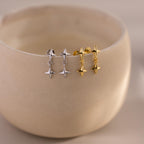 Two pairs of Star Drop Flatback Studs—celestial, star-shaped earrings in silver and gold—adorn the edge of a beige ceramic bowl. These versatile studs are perfect for cartilage piercings or as stylish drop earrings for any occasion.
