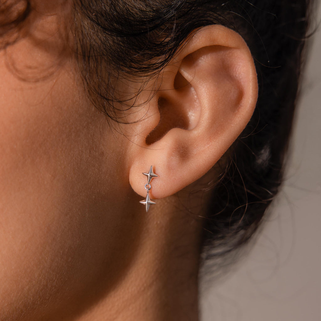 A woman’s ear wearing a silver flat back earring featuring two dangling star-shaped charms, subtly reflecting the light against her skin.